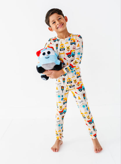 A young boy stands barefoot against a white studio background, wearing a long-sleeve pajama set with a colorful construction vehicle print and holding a plush toy.