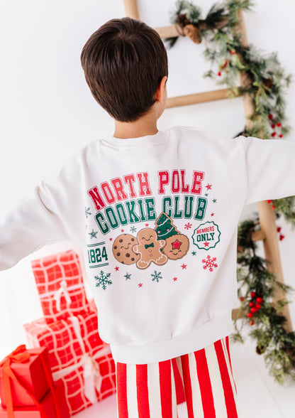 A child viewed from behind wearing a white sweatshirt with a colorful Christmas gingerbread theme graphic that reads 'NORTH POLE COOKIE CLUB' and 'MEMBERS ONLY', paired with red and white striped pants in a holiday decorated room.