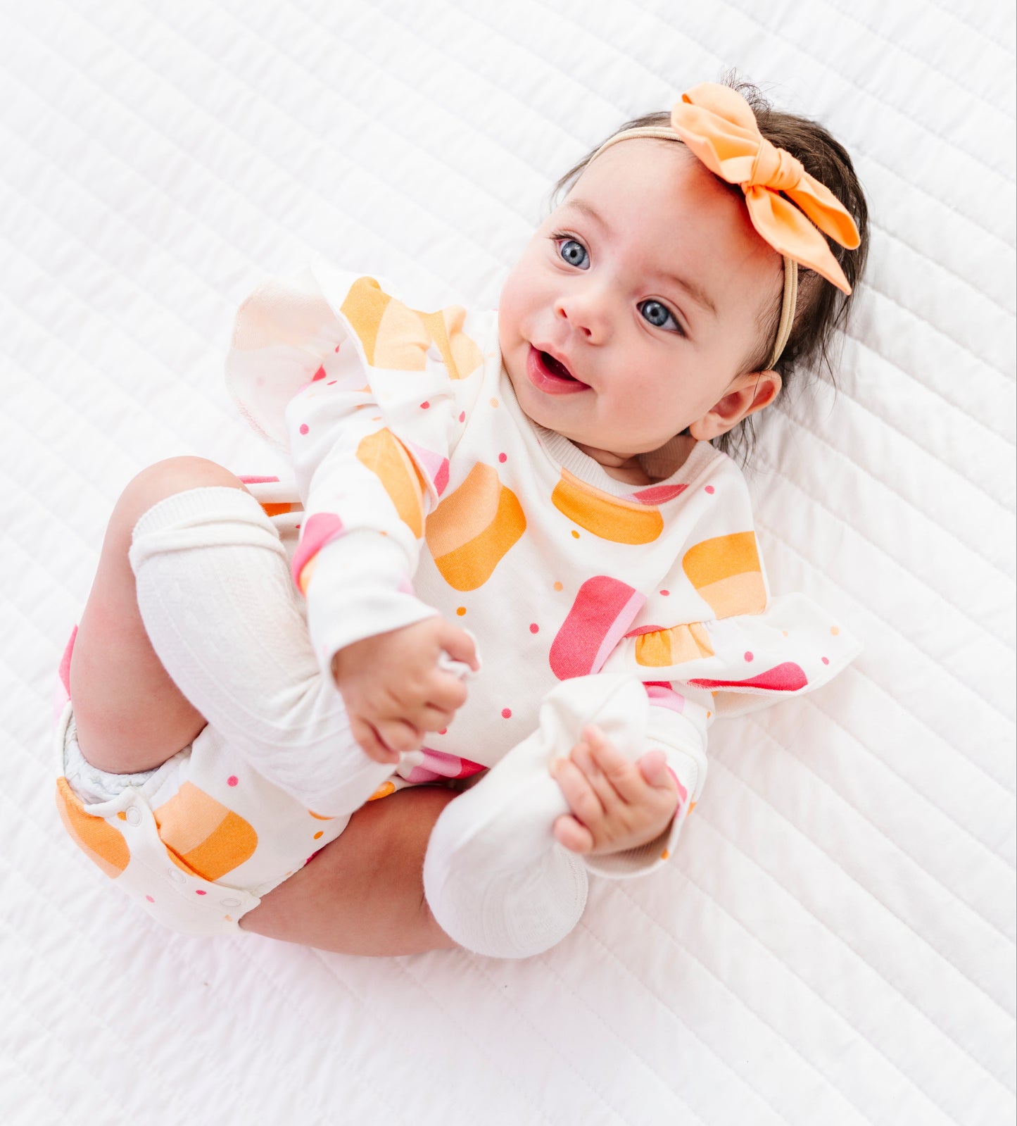 A baby lying on a white quilted blanket, wearing a white one-piece outfit with orange and pink ice cream cone patterns, white booties, and an orange bow headband.