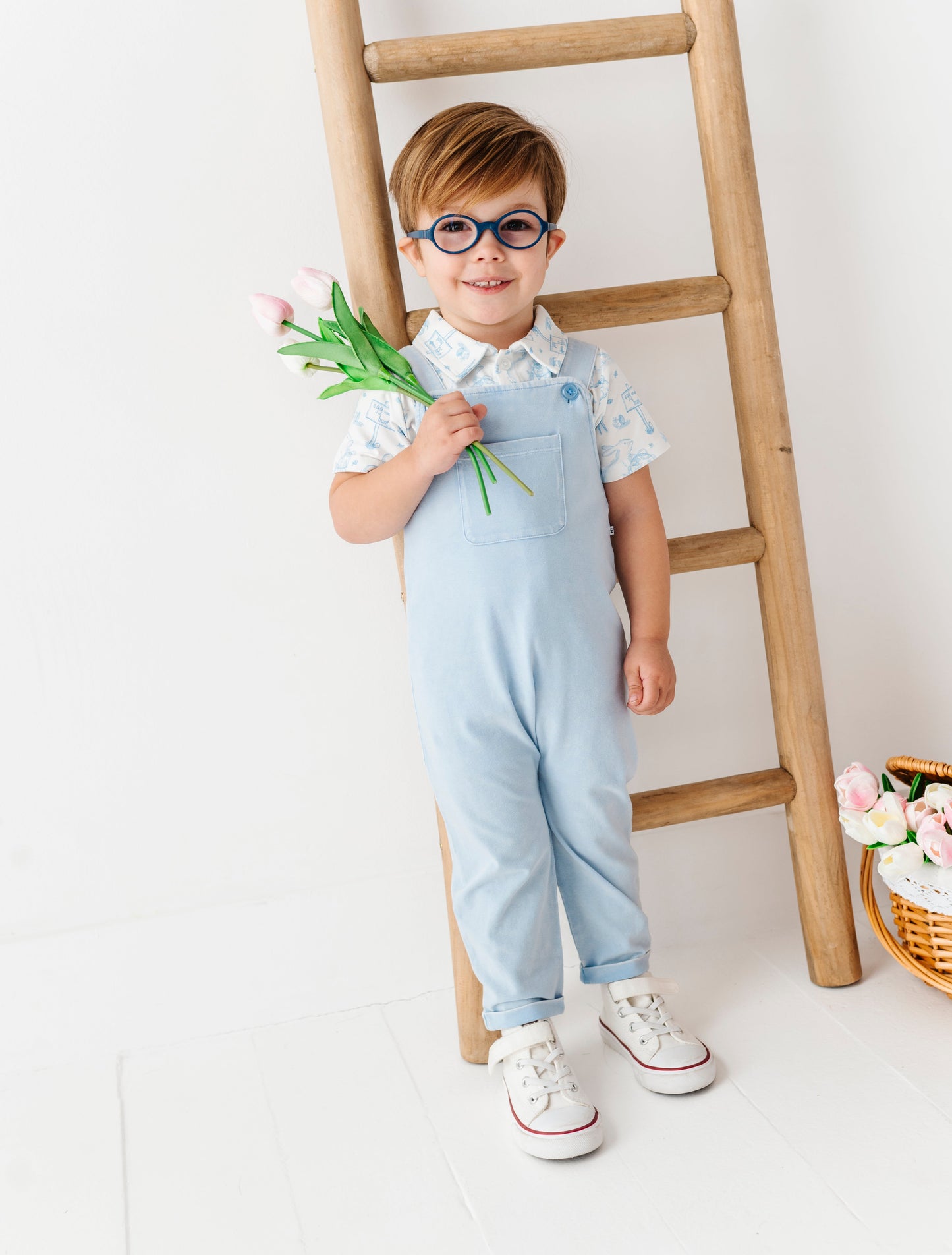 Child in light blue overalls holding flowers, standing next to a wooden ladder on a white background