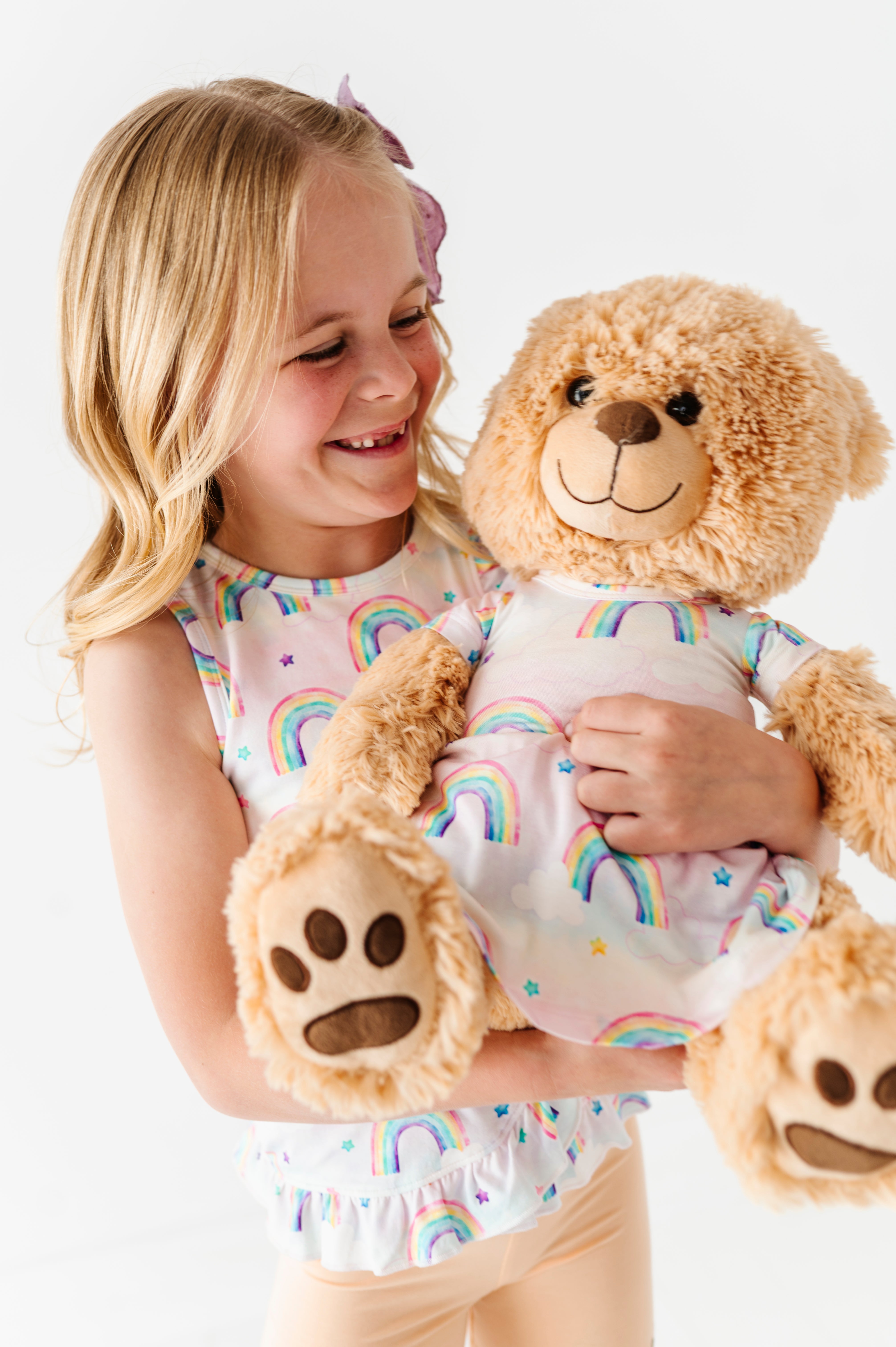 Little girl wearing a bamboo watercolor rainbows peplum top and holding a teddy bear wearing a bamboo dress in a matching rainbows print 