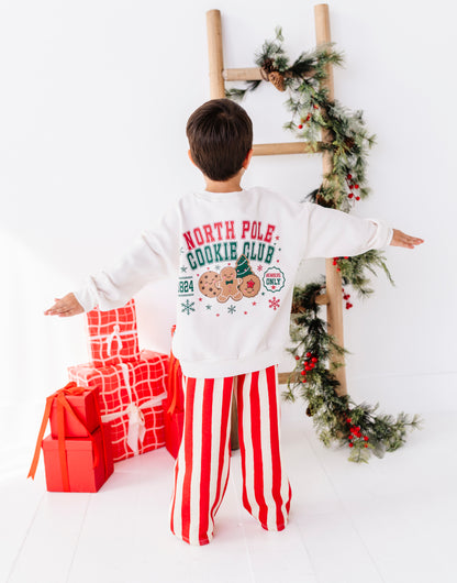 A child stands with arms outstretched, viewed from behind, wearing a white long-sleeve sweatshirt with a colorful 'North Pole Cookie Club' graphic and red-and-white vertically striped pants. The scene is decorated with wrapped red presents and a wooden ladder draped with green garland against a white wall.