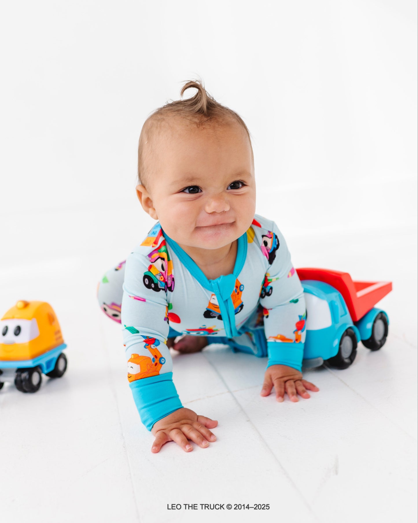 Baby in colorful Leo the Truck bamboo zip romper with Leo the Truck toys on a white background