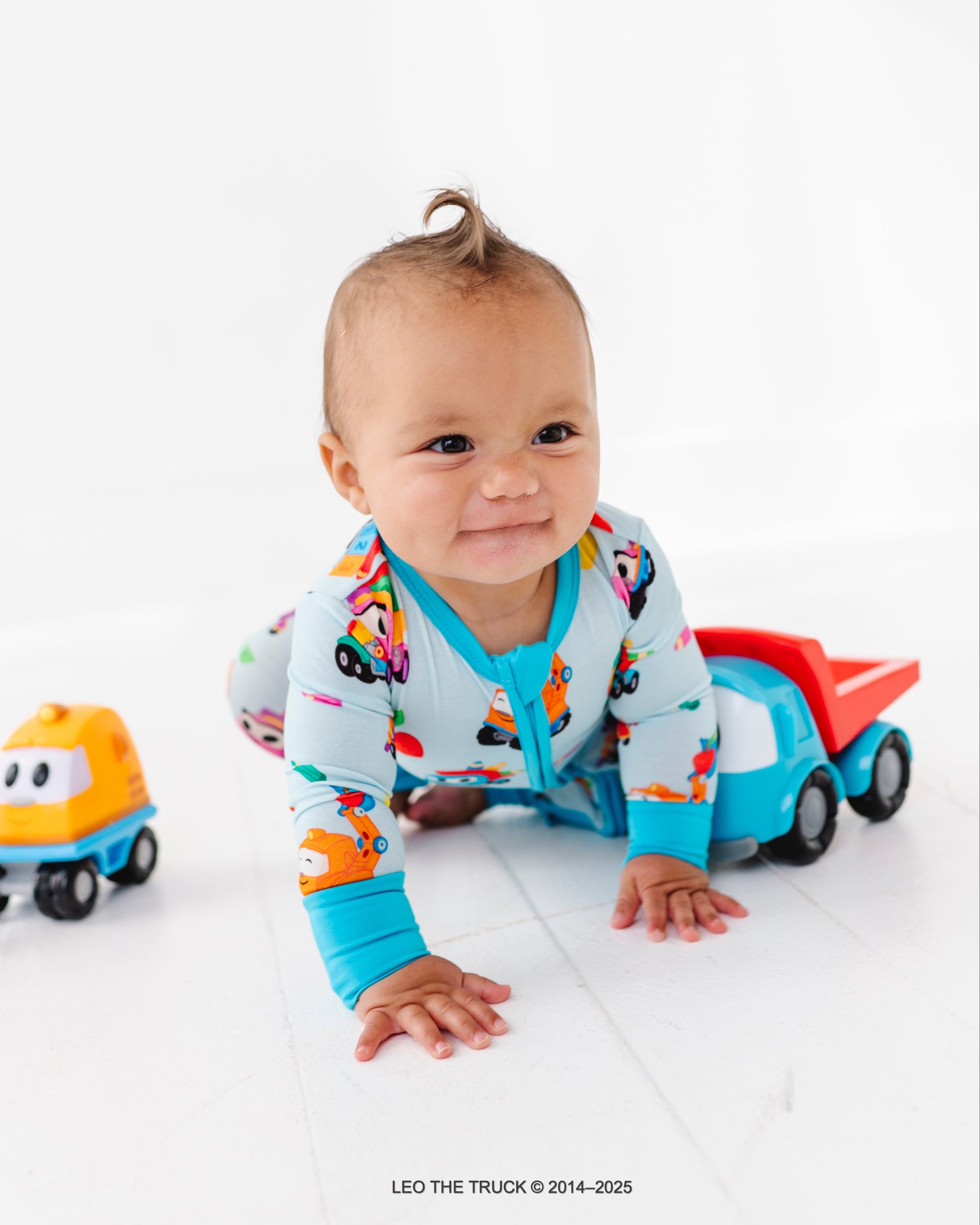 Baby in colorful Leo the Truck bamboo zip romper with Leo the Truck toys on a white background