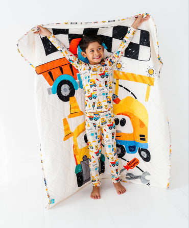 A child wearing a white one-piece pajama set with a colorful cartoon construction vehicle print stands barefoot and holds up a matching quilted blanket with a large construction truck and yellow crane design, set against a plain white studio background.