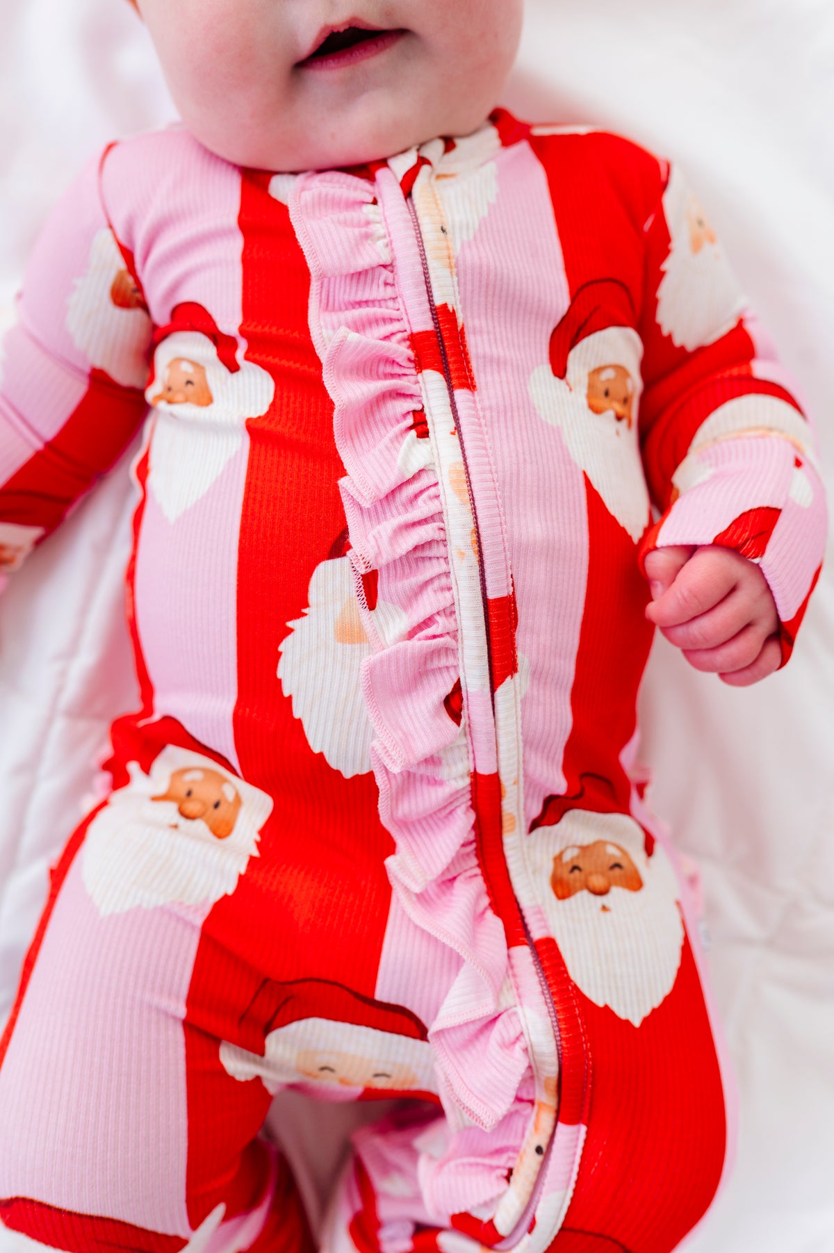 A close-up of a baby wearing a red and pink striped ribbed one-piece bamboo sleeper with ruffle trim and Santa Claus face prints, lying on a white blanket.