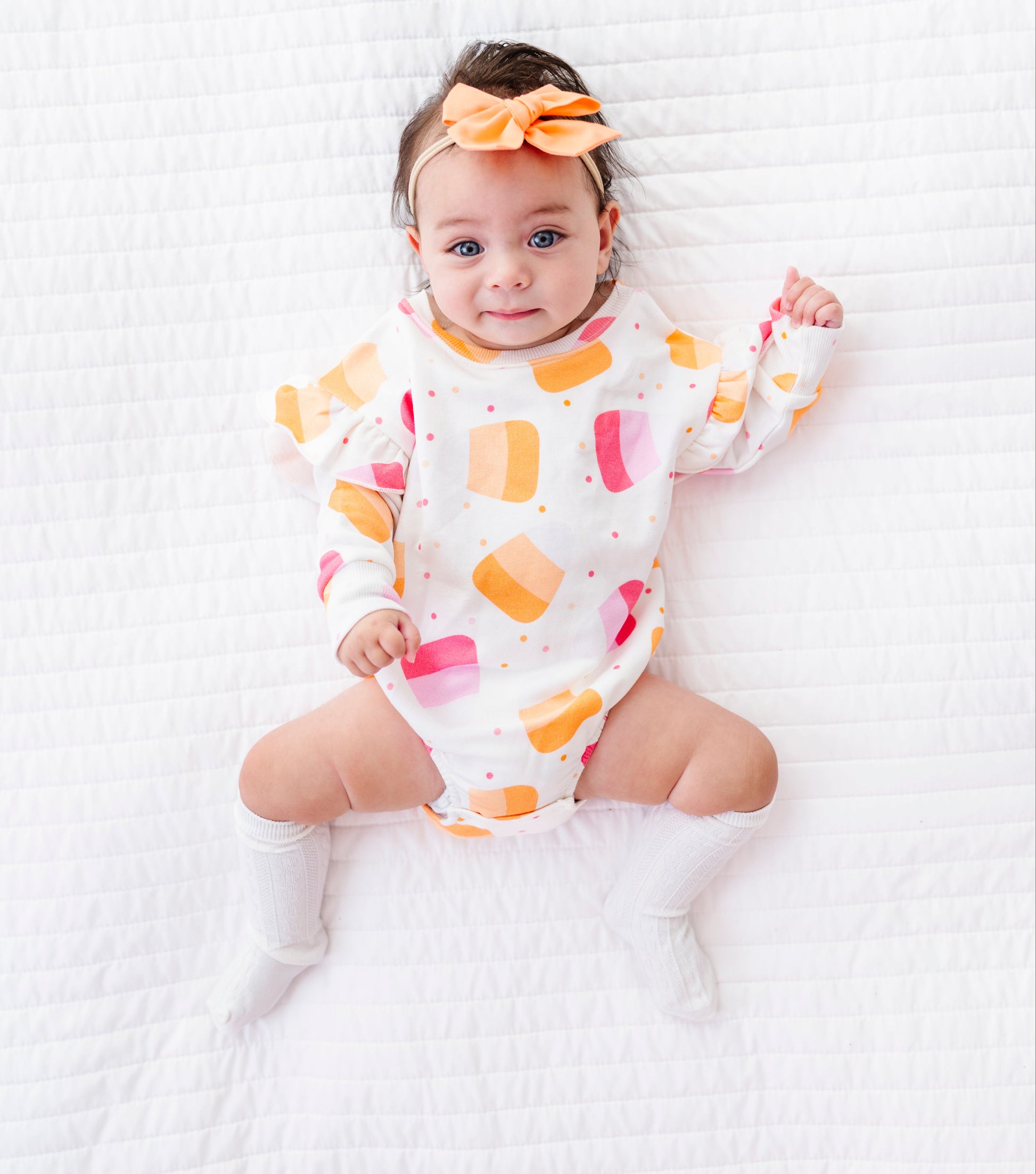 A baby lying on a white quilted blanket, wearing a white long-sleeve romper with a colorful ice cream cone pattern in orange, pink, and yellow, along with a matching orange bow headband and white socks.