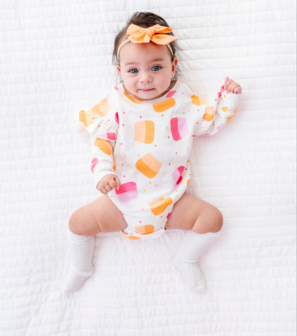 A baby lying on a white quilted blanket, wearing a white long-sleeve romper with a colorful ice cream cone pattern in orange, pink, and yellow, along with a matching orange bow headband and white socks.