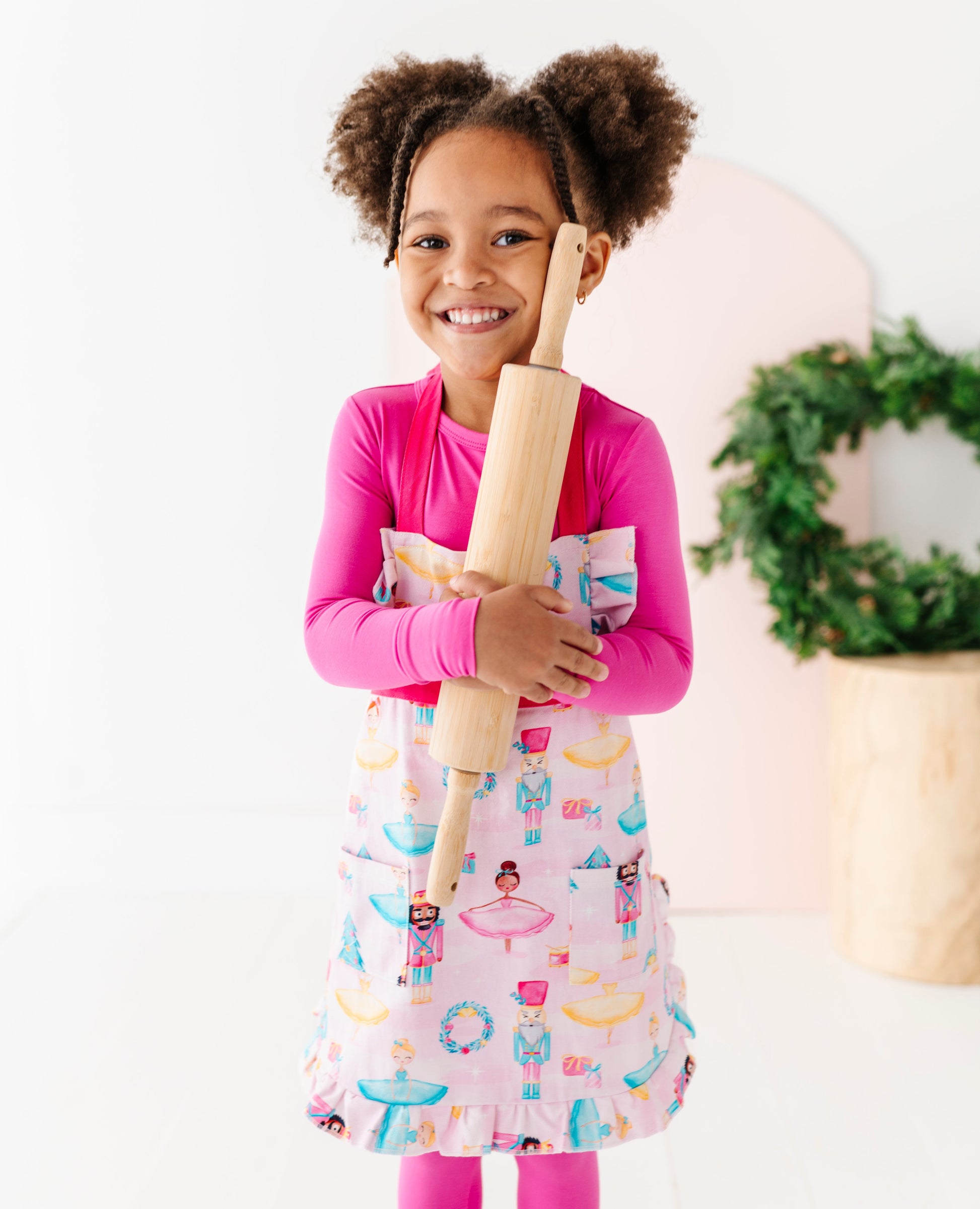 Child in a pink shirt and colorful sugarplum apron holding a rolling pin, standing against a white background.