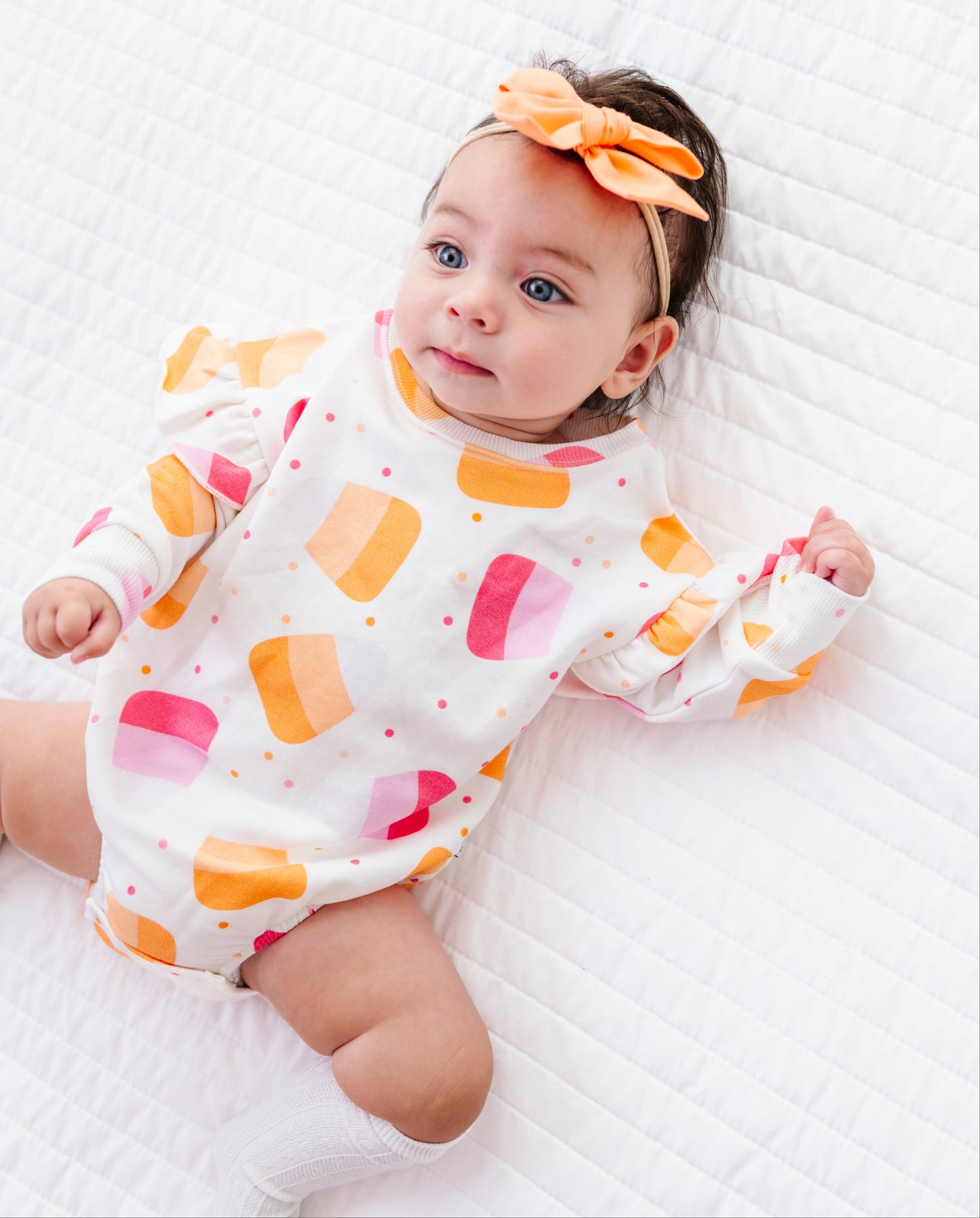 A baby lying on a white quilted blanket, wearing a white long-sleeve romper with a colorful ice cream cone pattern in orange, pink, and yellow, along with an orange bow headband.