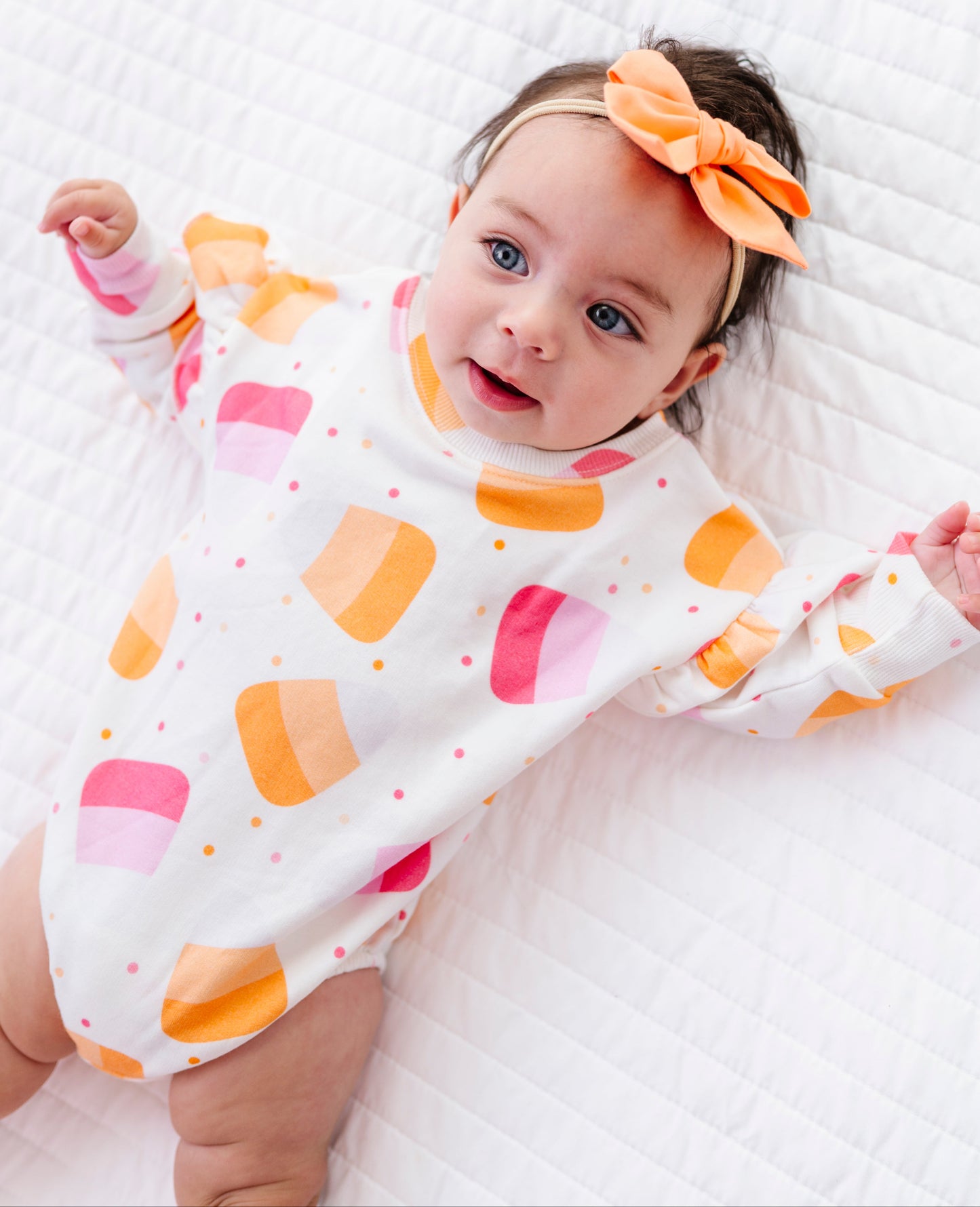 A baby lying on a white quilted blanket, wearing a white long-sleeve bodysuit with pink and orange geometric shapes, a peach-orange bow headband, and smiling.