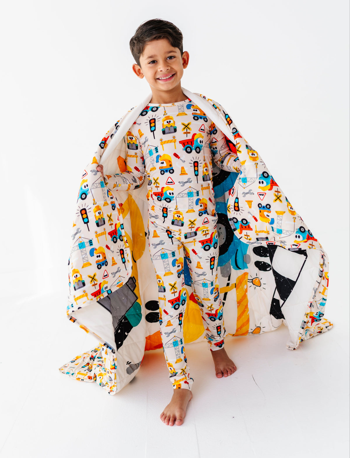 A young boy stands barefoot on a white studio floor, wearing a matching two-piece pajama set and holding a large quilted blanket with a colorful construction vehicle print.