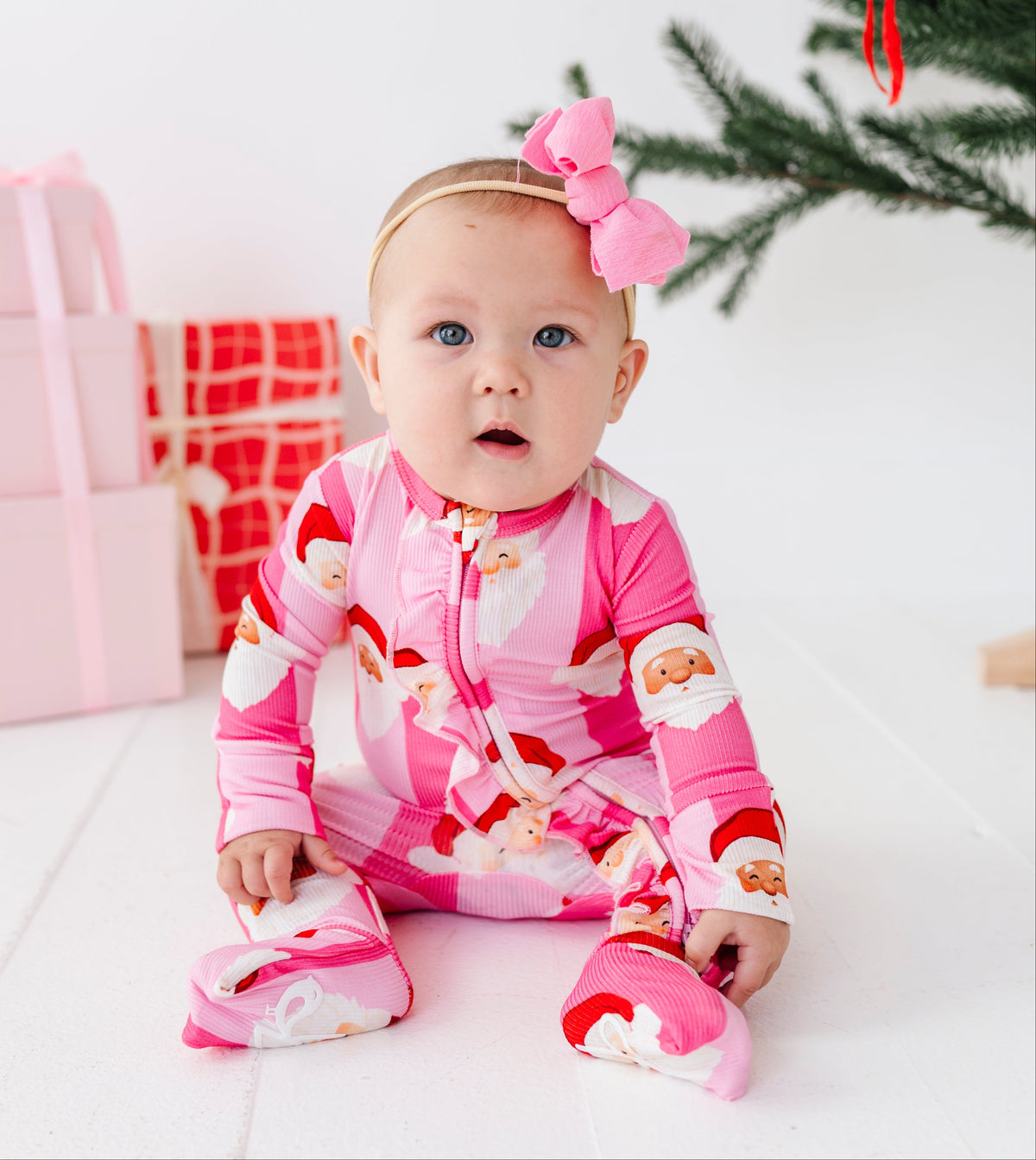 A baby wearing a pink footed bamboo convertible romper with a Santa Claus pattern and a matching pink bow headband, sitting on a white floor next to wrapped gifts and a Christmas tree.