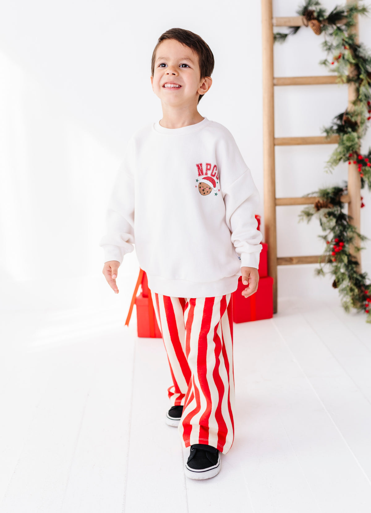 A young boy smiling and walking while wearing a white crewneck sweatshirt with a red and white holiday graphic on the chest, paired with red and white vertically striped pants and black slip-on shoes. The background is a bright white studio setting with a wooden ladder and Christmas garland.