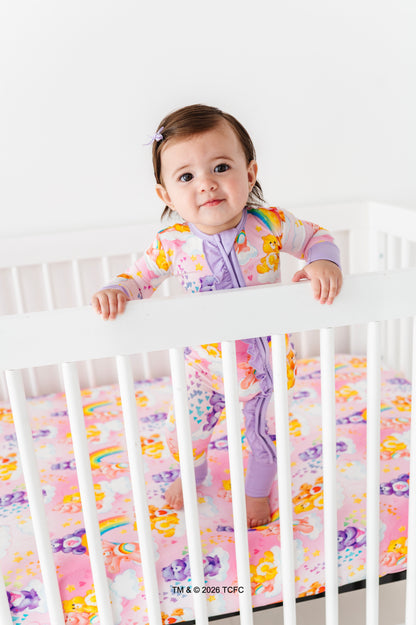 Baby in a colorful Care Bears™ zip romper standing in a crib with a white background