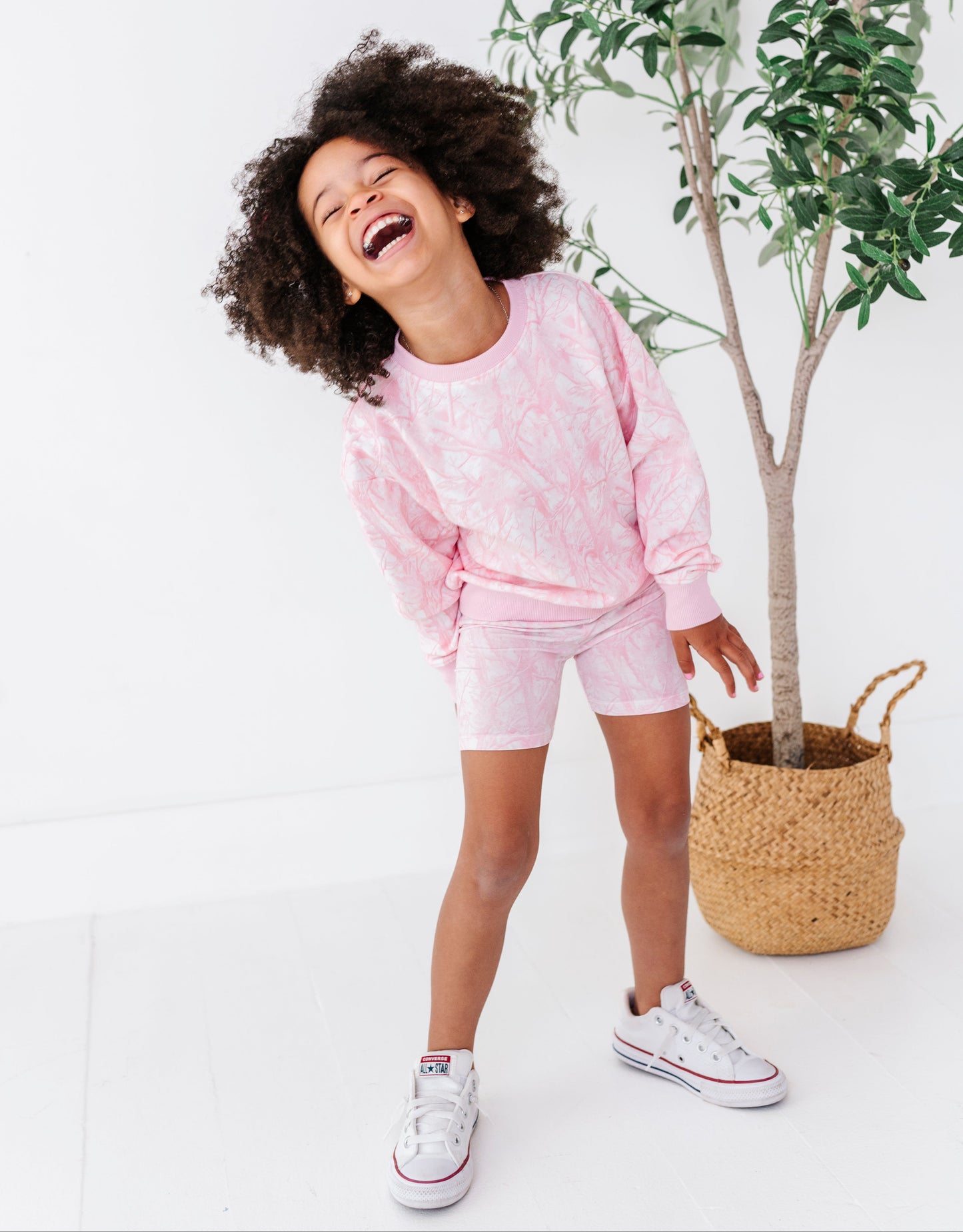 Child in pink camo outfit standing next to a plant on a white background