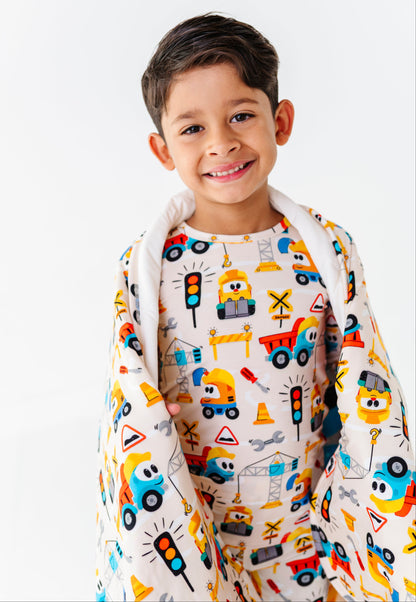 A young boy smiling while wearing a white long-sleeve pajama shirt and matching blanket set with a colorful construction vehicle pattern, set against a plain white background.