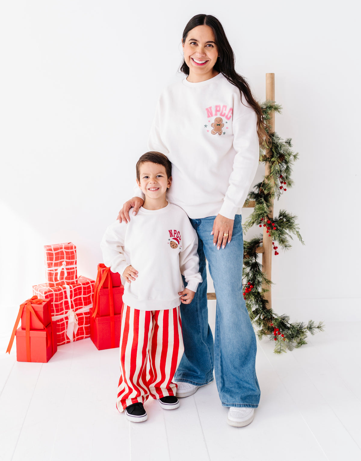 A woman and a young boy wearing matching white holiday sweatpants and sweatshirts with red and pink Christmas graphics, standing in a bright studio with wrapped presents and a Christmas garland.