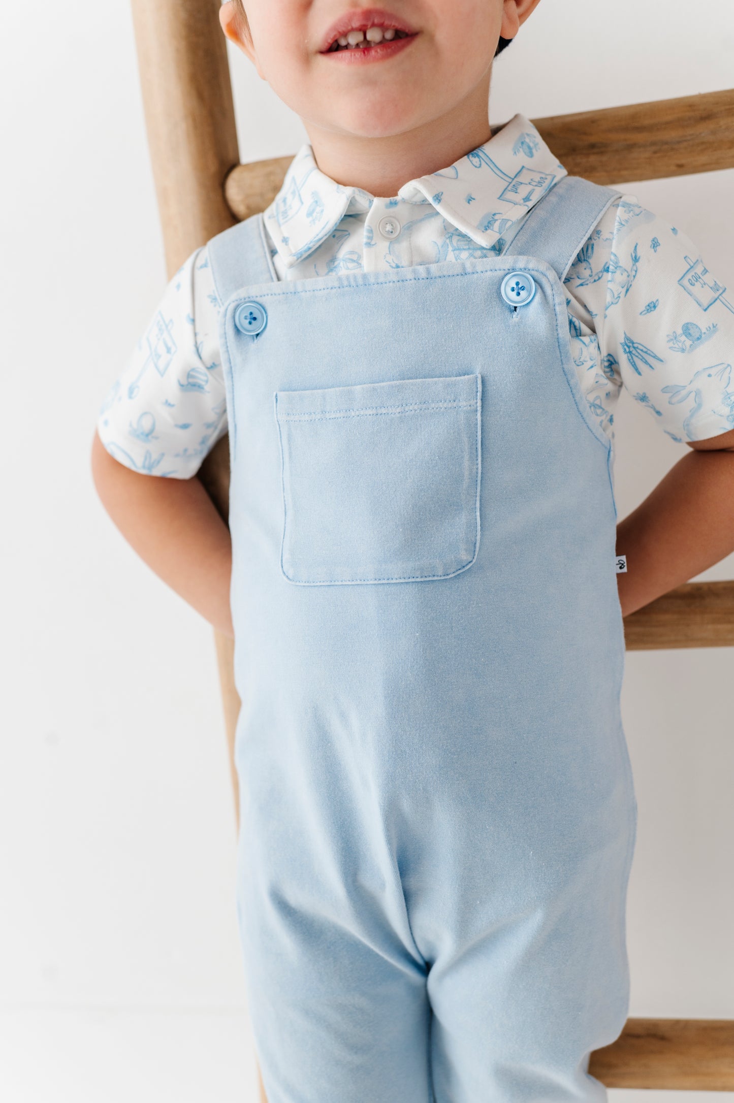 Child wearing light blue overalls and a patterned Easter shirt against a white background