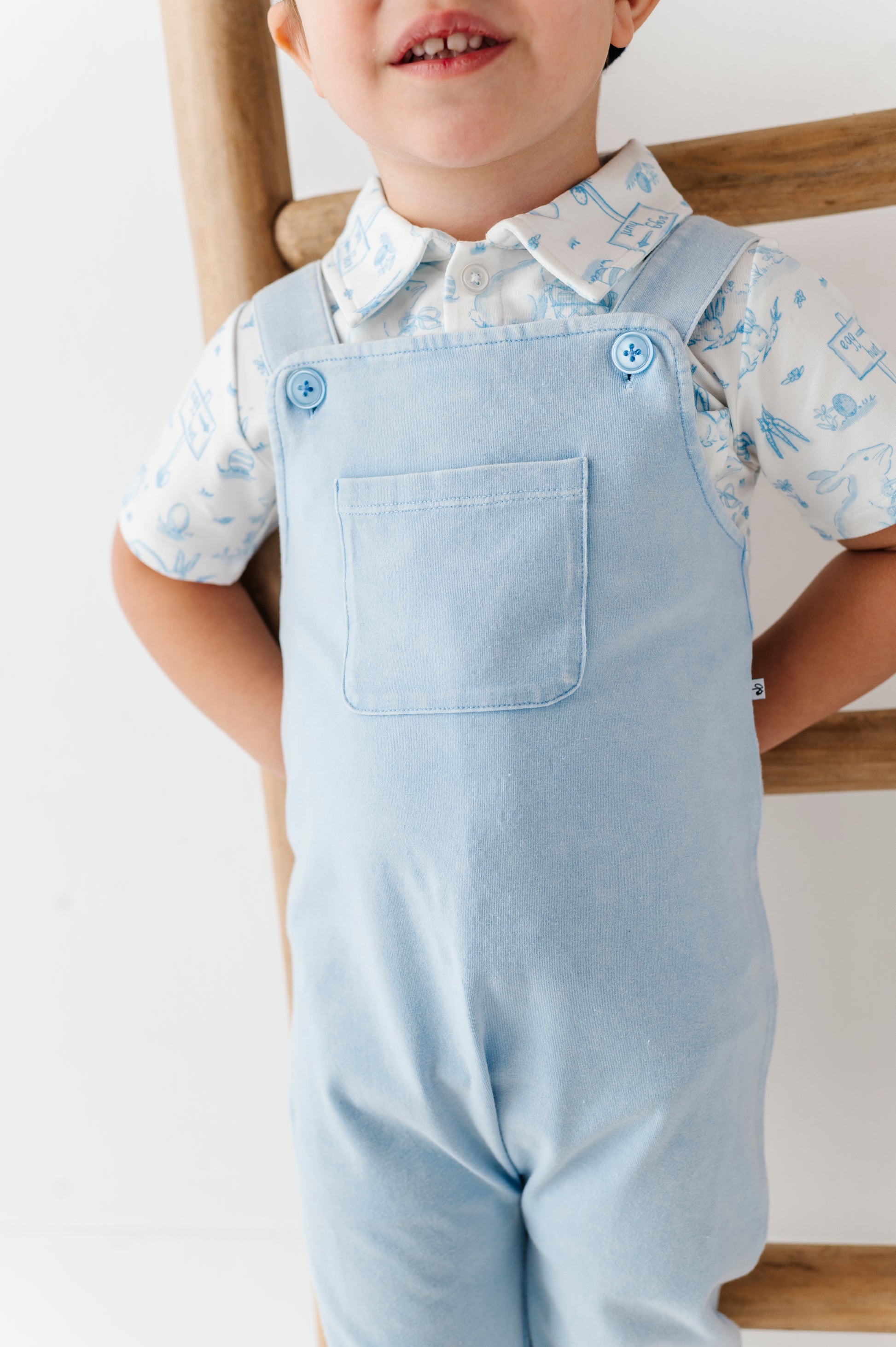 Child wearing light blue overalls and a patterned Easter shirt against a white background