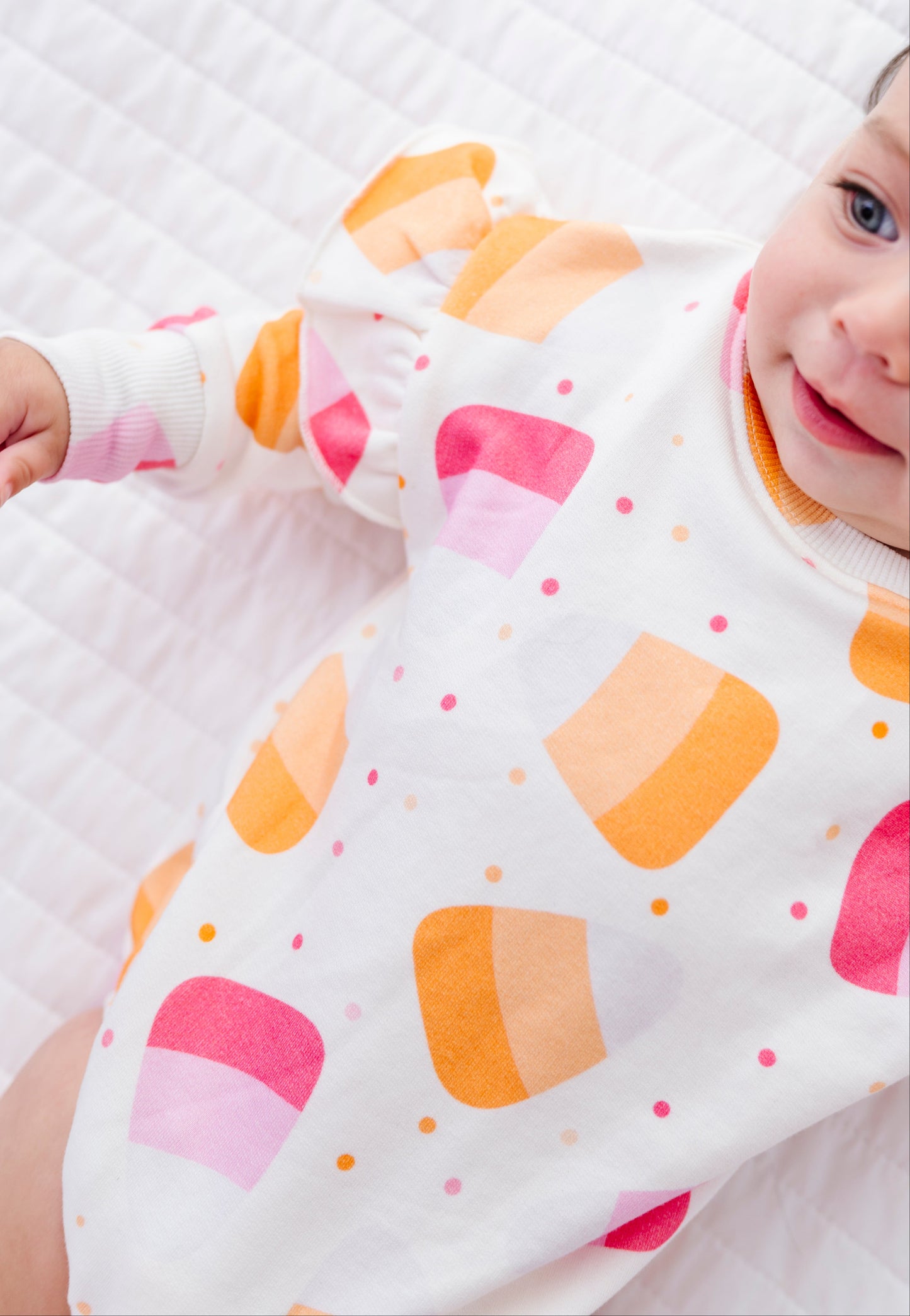 A baby lying on a white quilted blanket, wearing a white long-sleeve bodysuit with a colorful pattern of pink and orange half-cupcake shapes and small pink and orange dots.