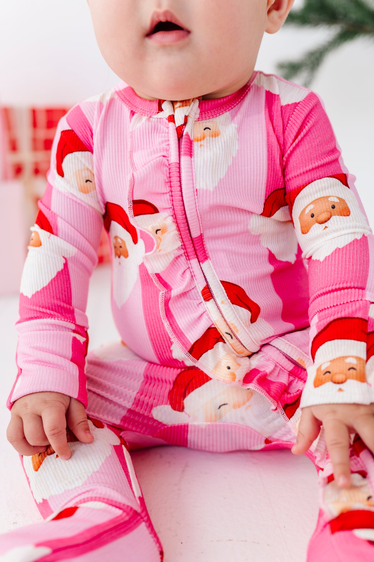 A baby wearing a pink ribbed zip-up bamboo sleeper with a Santa Claus print, sitting on a pink surface.