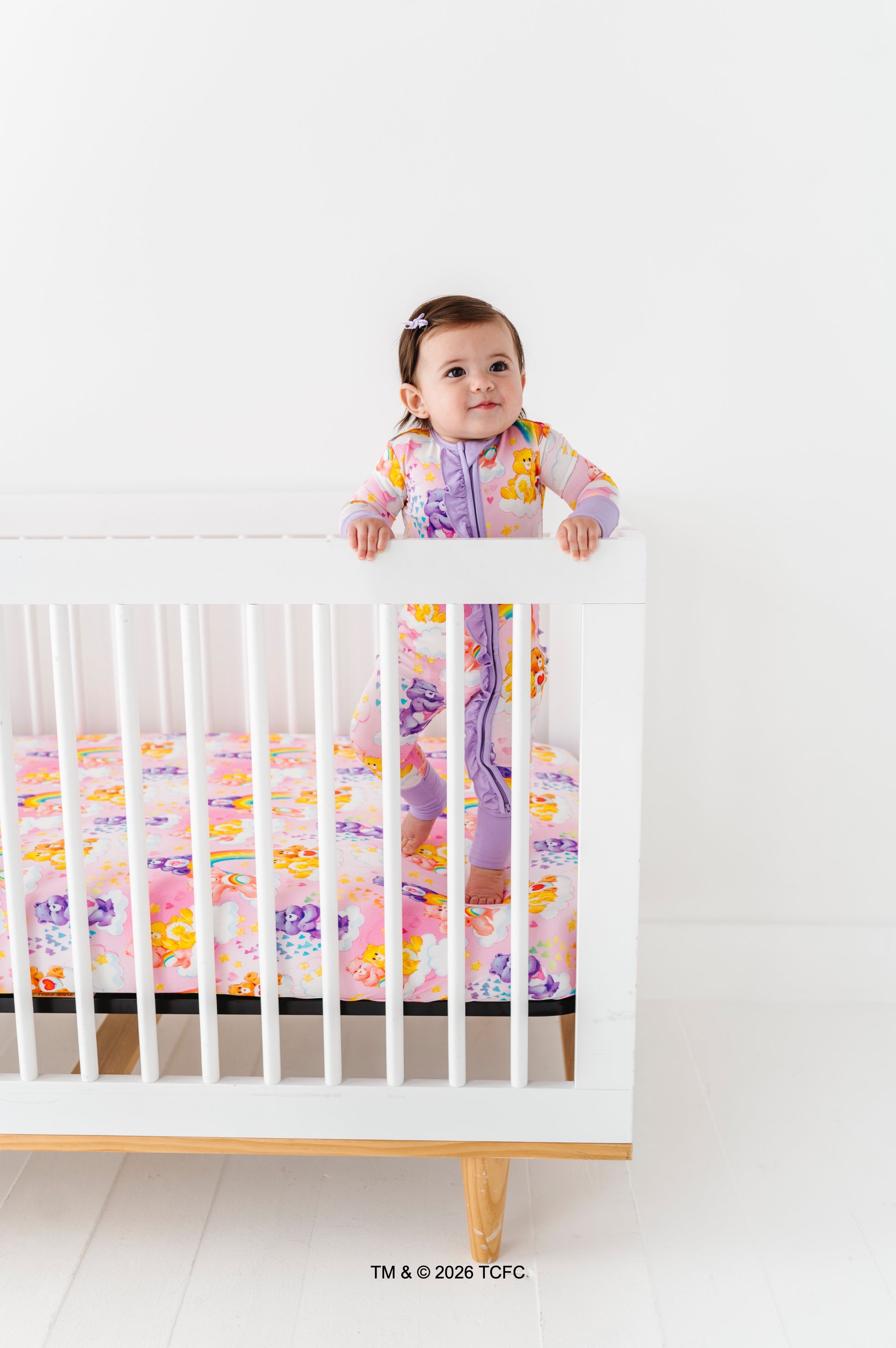 Baby standing on a crib with colorful Care Bears™ bedding against a white background