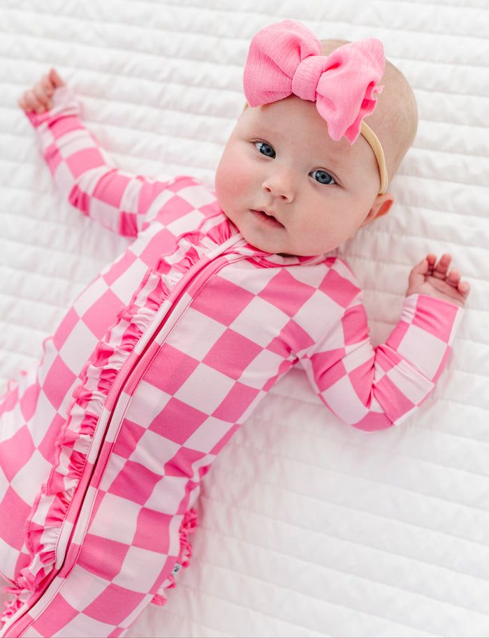 Baby in pink checkered bamboo convertible romper with a pink bow on a white textured background