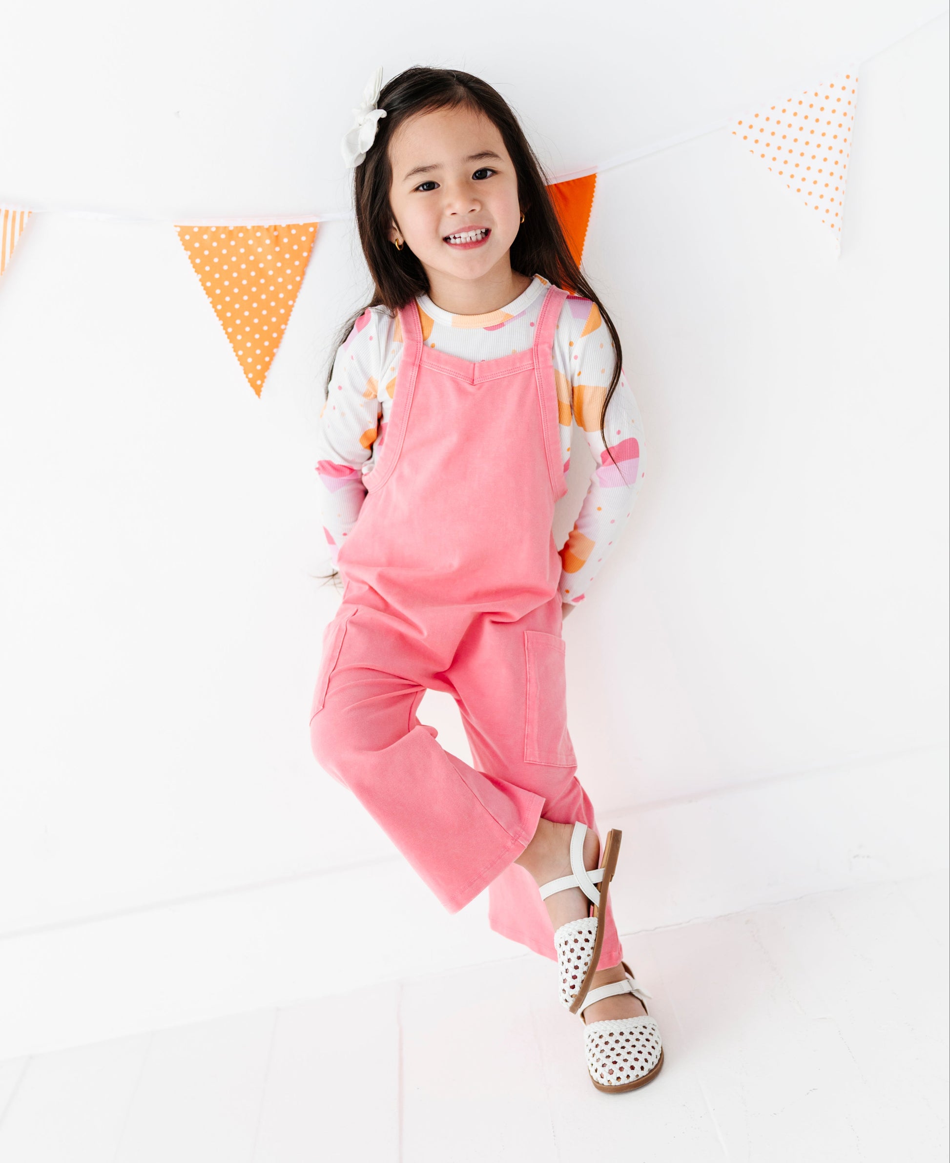 A young girl smiling and posing in a pink overall jumpsuit over a white long-sleeve shirt with orange and pink patterns, wearing white polka-dot sandals, set against a white wall with orange and white polka dot bunting.