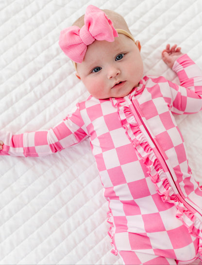 Baby in pink checkered bamboo convertible romper with a pink bow on a white textured surface