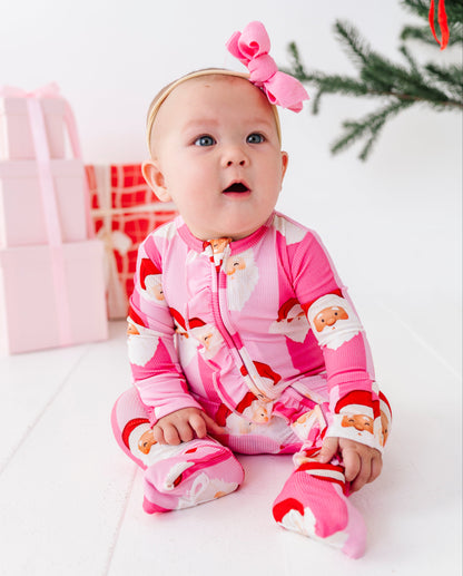 A baby sitting on a white floor wearing a pink Santa Claus print footed bamboo sleeper and a matching pink bow headband, with gift boxes and a Christmas tree branch in the background.