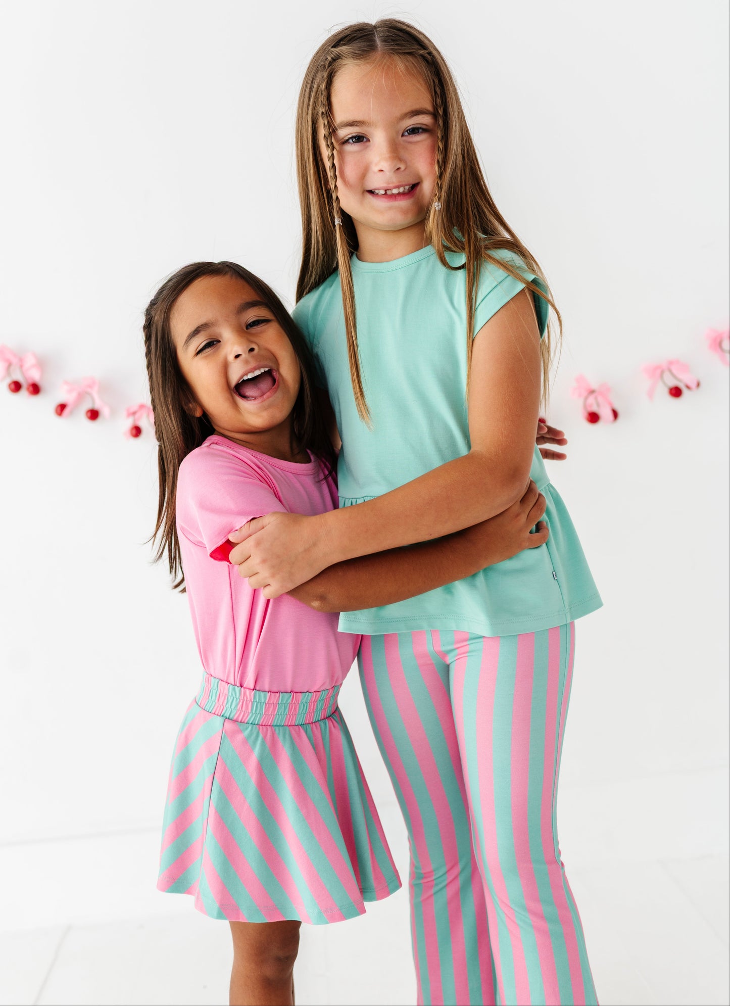 Two young girls hugging in a bright studio setting. The girl on the left wears a pink long-sleeve top and a pink and mint green striped skirt, while the girl on the right wears a mint green short-sleeve top and matching pink and mint green striped pants.