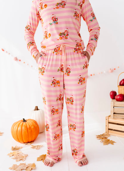 A person wearing a pink long-sleeve pajama set with horizontal stripes and a small floral print, standing barefoot in a styled indoor scene with pumpkins, fall leaves, and a basket of red apples.