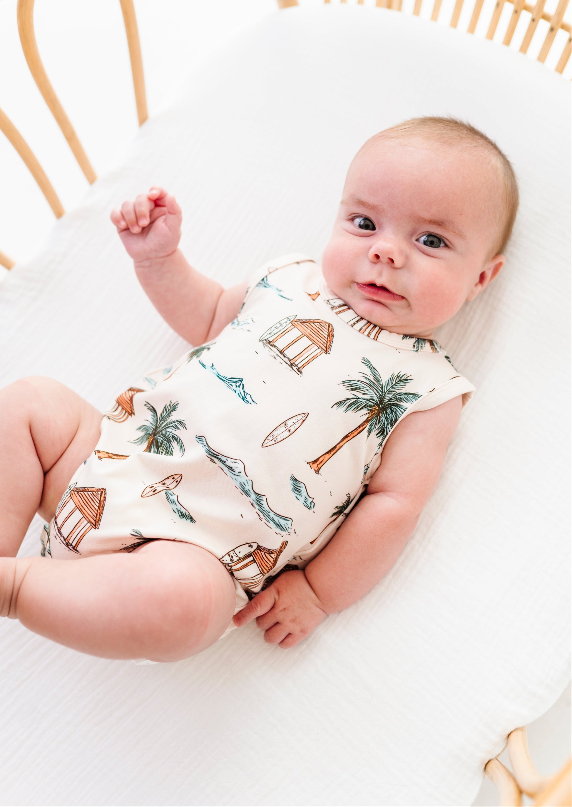 Baby lying on a white bassinet wearing a sleeveless romper with beach-themed design.