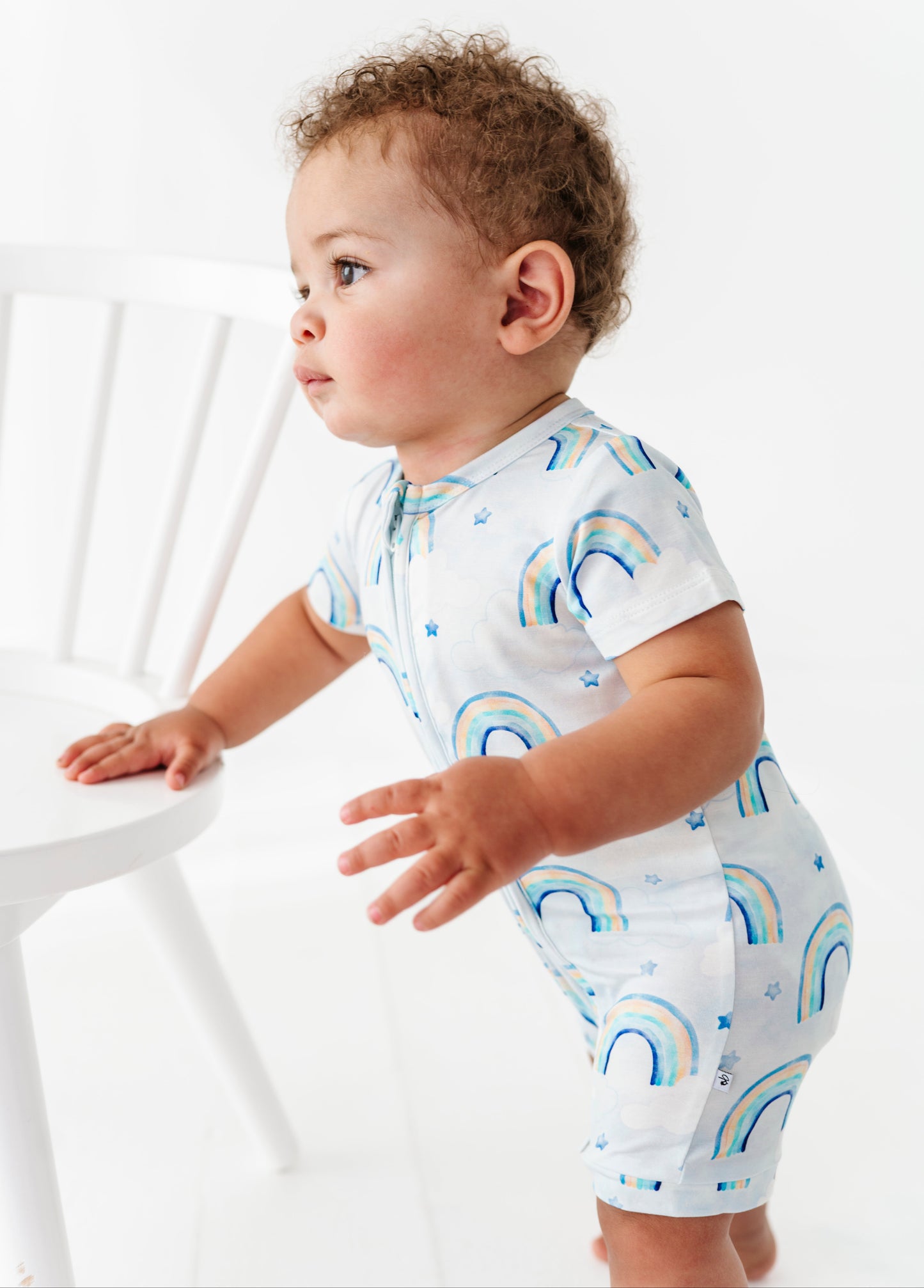 Baby wearing a light blue bamboo shortie romper with blue watercolor rainbow patterns on a white background