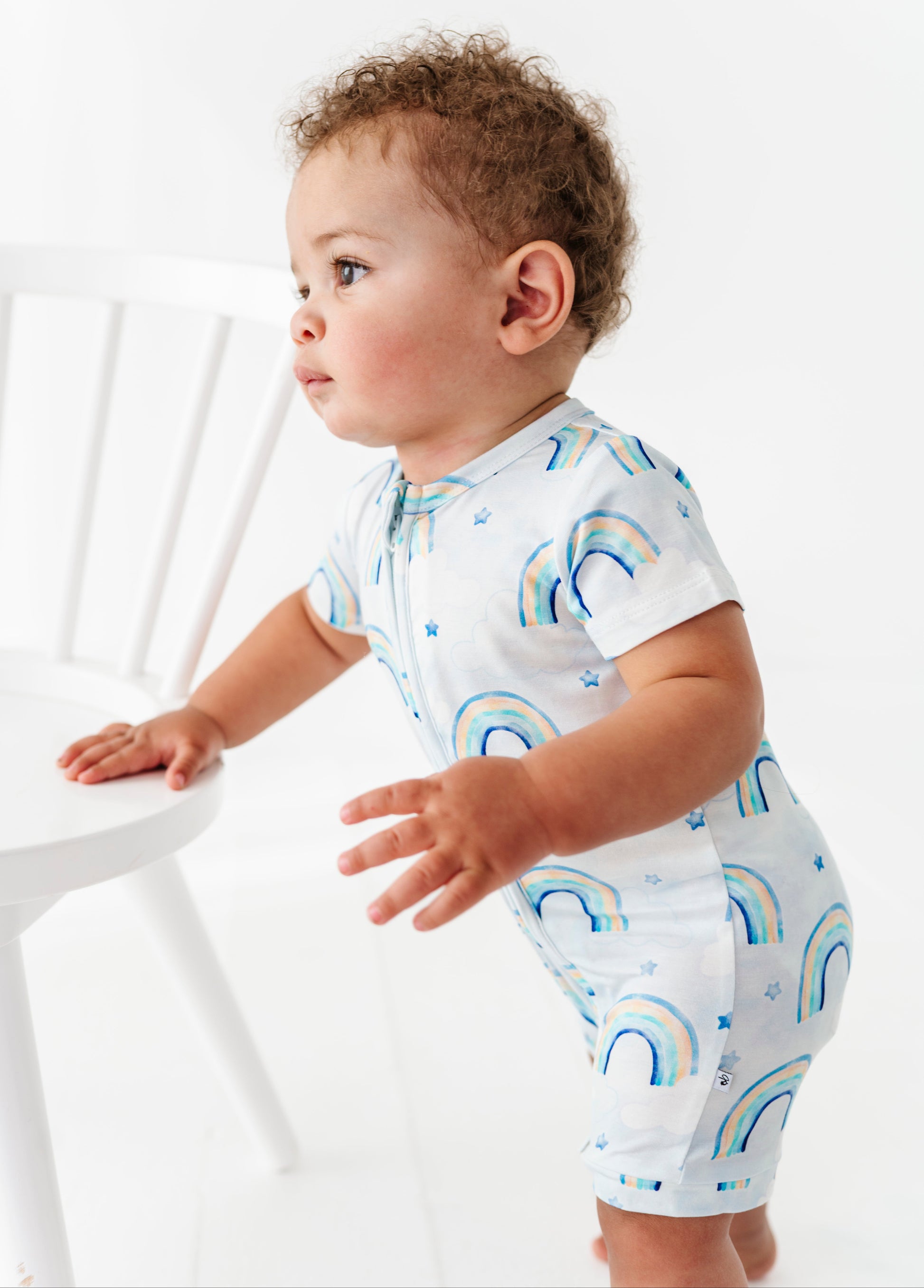Baby wearing a light blue bamboo shortie romper with blue watercolor rainbow patterns on a white background
