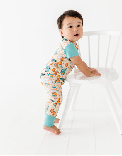 Baby in a patterned sports bamboo zip romper standing by a white chair against a white background