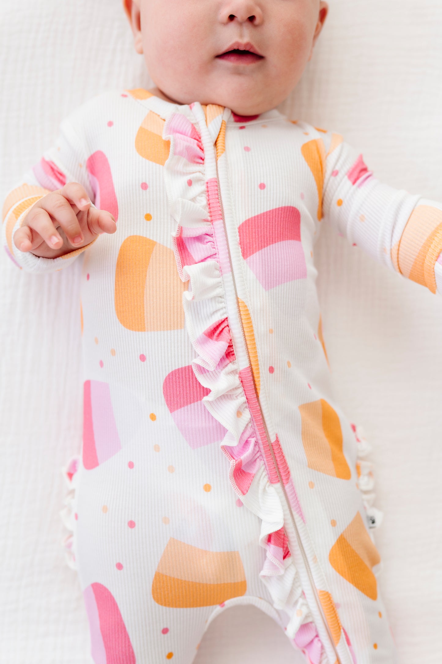 A close-up of a baby wearing a white long-sleeve footless sleeper with pink and orange geometric shapes, ruffle trim, and a front zipper, lying on a white textured blanket.