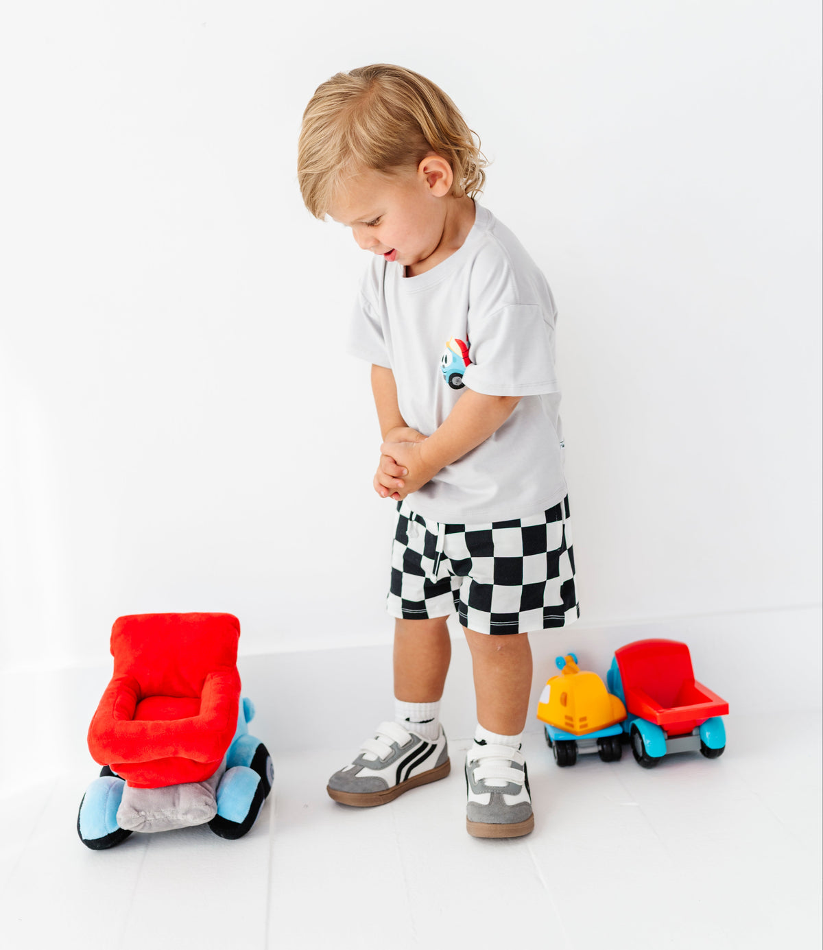 A toddler stands on a white floor wearing a white t-shirt and black-and-white checkered shorts, looking down toward two small toy vehicles on a white studio background.