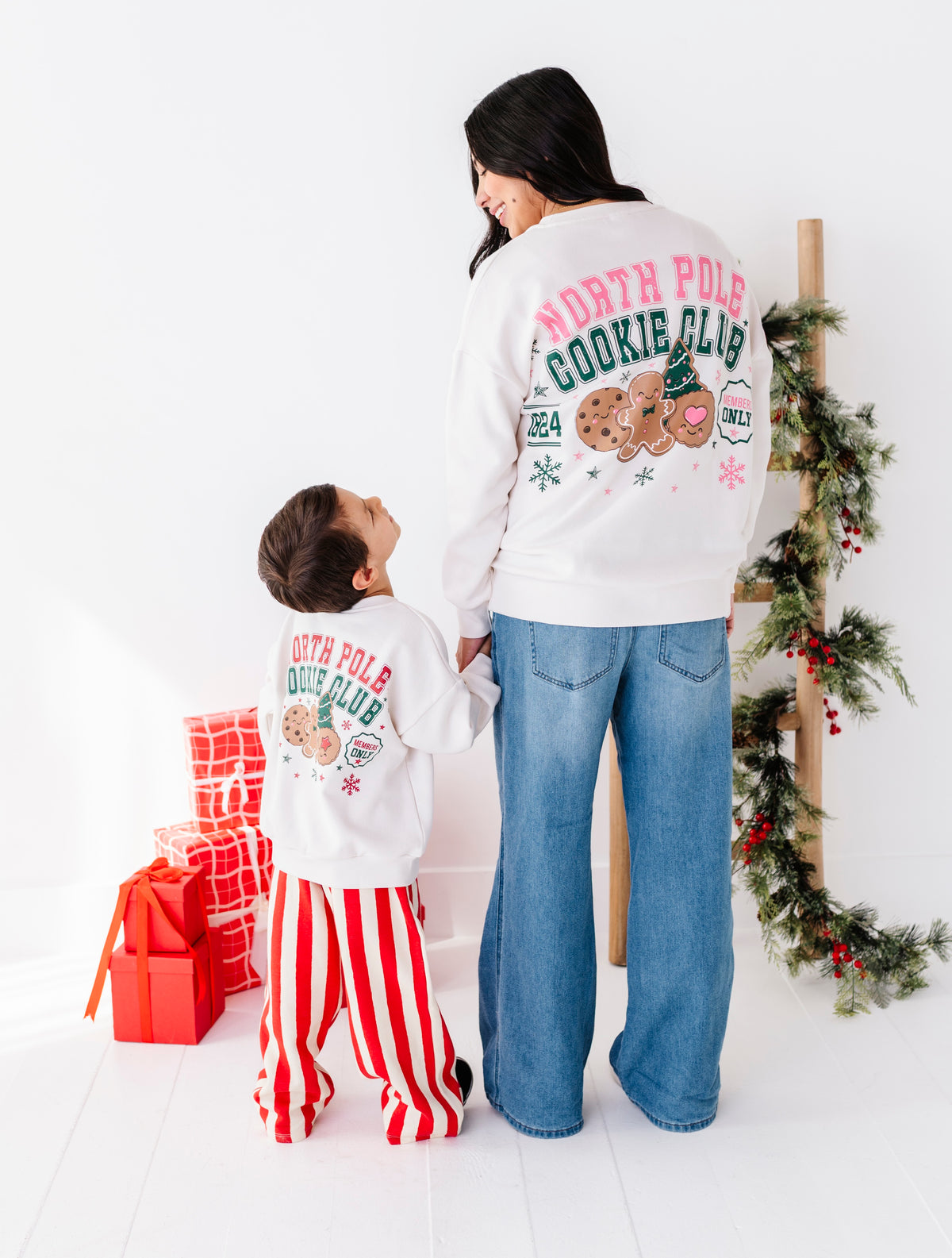 A woman and a child wearing matching white 'North Pole Cookie Club' sweatshirts with colorful holiday graphics, standing in a bright room with Christmas decorations and wrapped gifts.