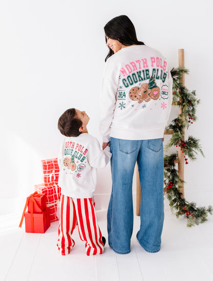 A woman and a child wearing matching white 'North Pole Cookie Club' sweatshirts with colorful holiday graphics, standing in a bright room with Christmas decorations and wrapped gifts.
