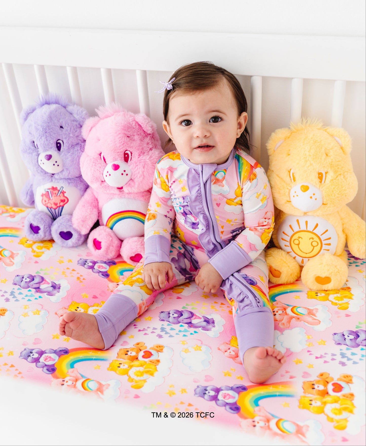 Baby in a colorful outfit sitting on a bed with Care Bears toys.