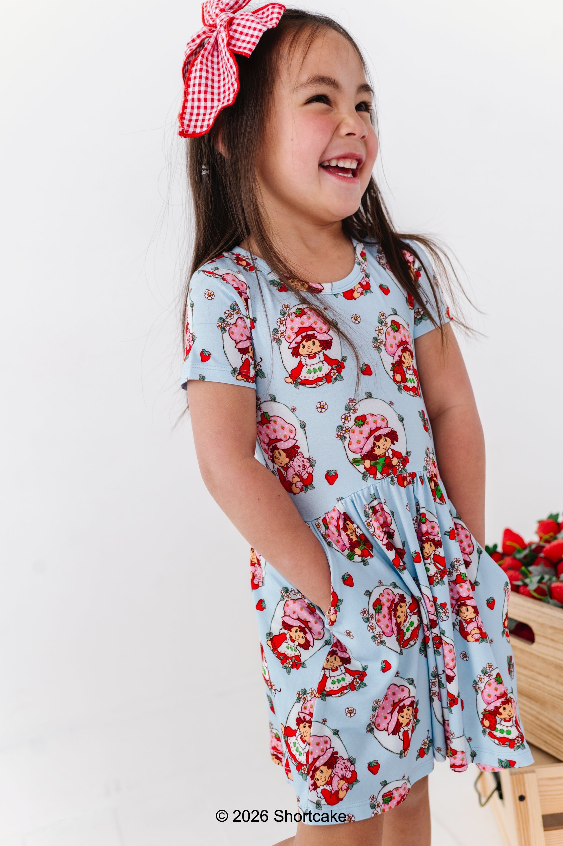Young girl wearing a Strawberry Shortcake™ dress with a red bow, standing against a white background.