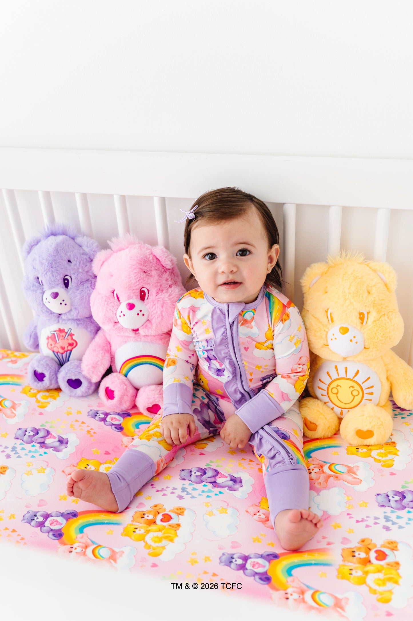 Baby in a colorful Care Bears™ zip romper sitting in a crib with matching bamboo crib sheet