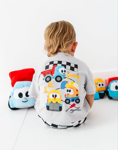 A young child with blonde hair sits with their back to the camera, wearing a white t-shirt with a colorful cartoon construction vehicle graphic on the back. The child is surrounded by plush toy vehicles on a clean white floor against a white wall.