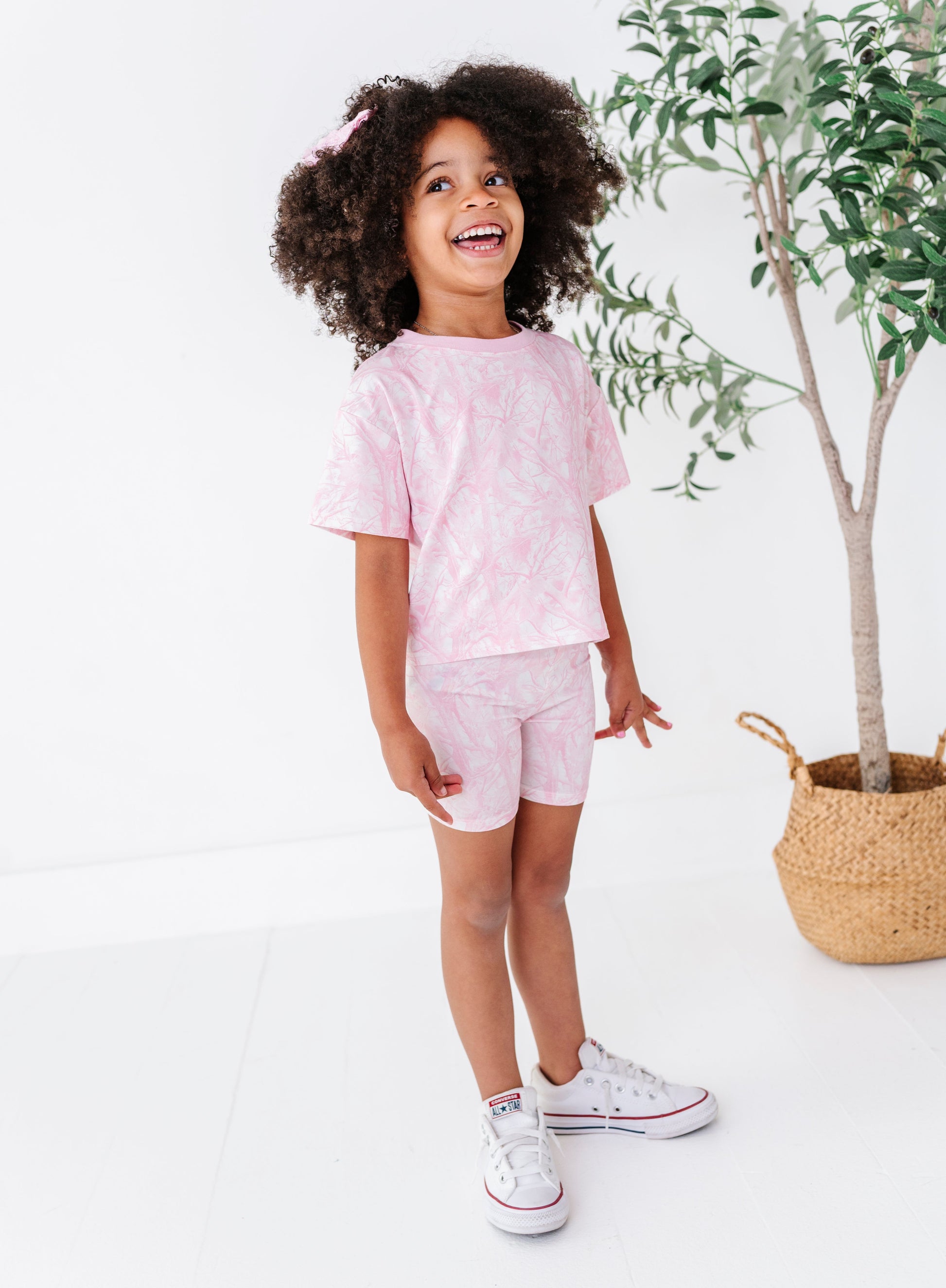Child wearing a pink outfit pink tree camo standing in a bright room with a plant in the background.