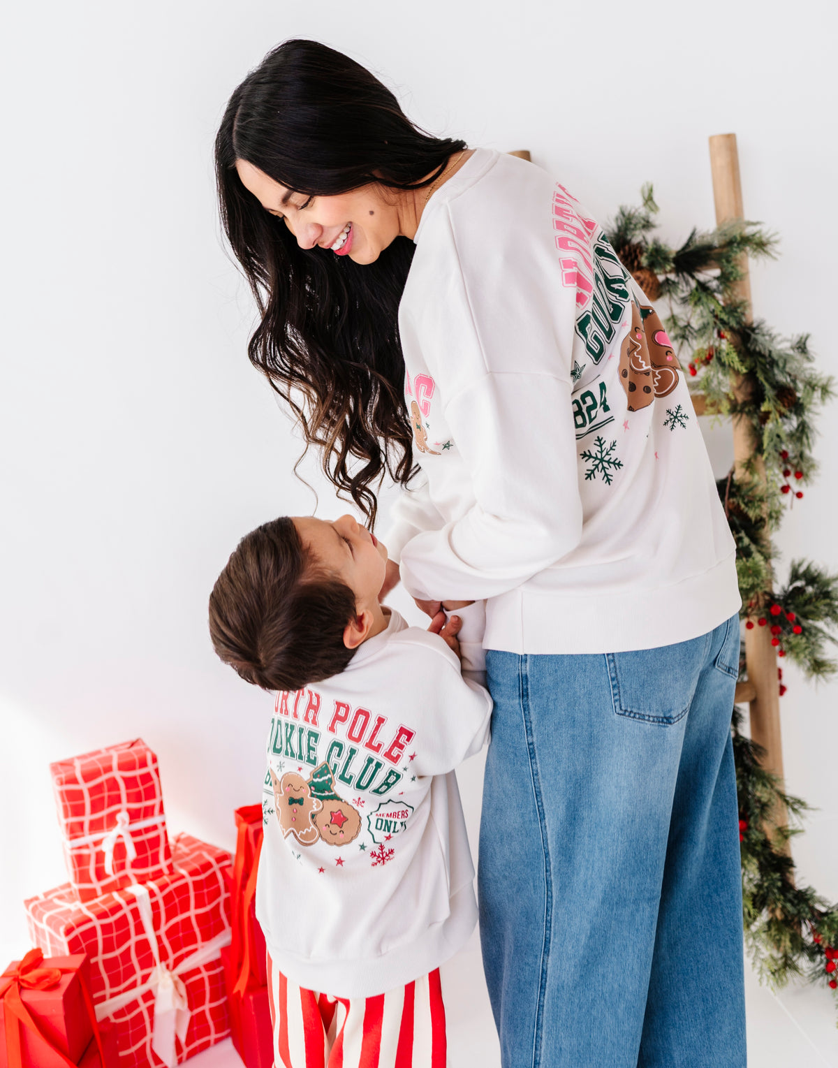A woman and a young boy wearing matching white Christmas-themed sweatshirts with colorful graphics, standing in a festive indoor setting with wrapped gifts and a decorated tree.