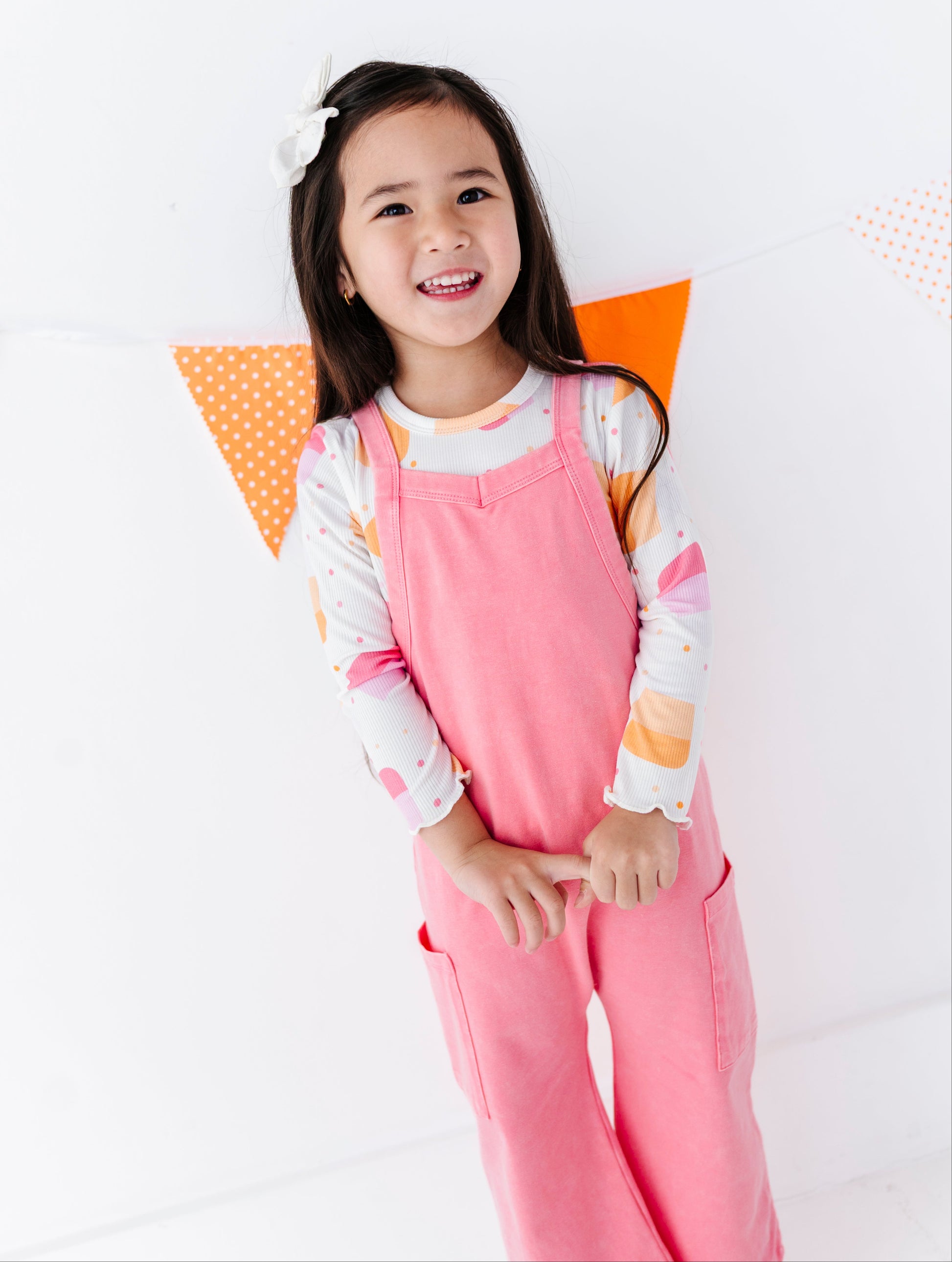 A young girl smiling and posing in a pink overall jumpsuit over a long-sleeve white shirt with pastel orange and pink patterns. She is standing against a white wall with orange and white bunting.