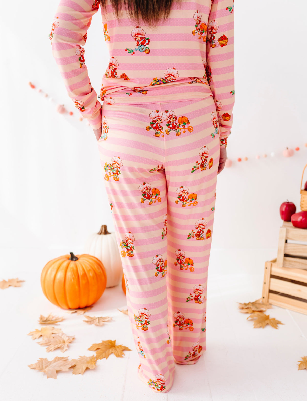 A person wearing a pink striped pajama set with a small floral and pumpkin print, shown from the back in a fall-themed studio setting with pumpkins, leaves, and wooden crates.