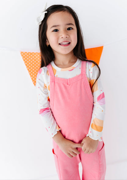 A young girl smiling and wearing a pink overall jumpsuit over a white long-sleeve shirt with pastel orange and pink patterns, standing against a white wall with orange polka dot bunting.